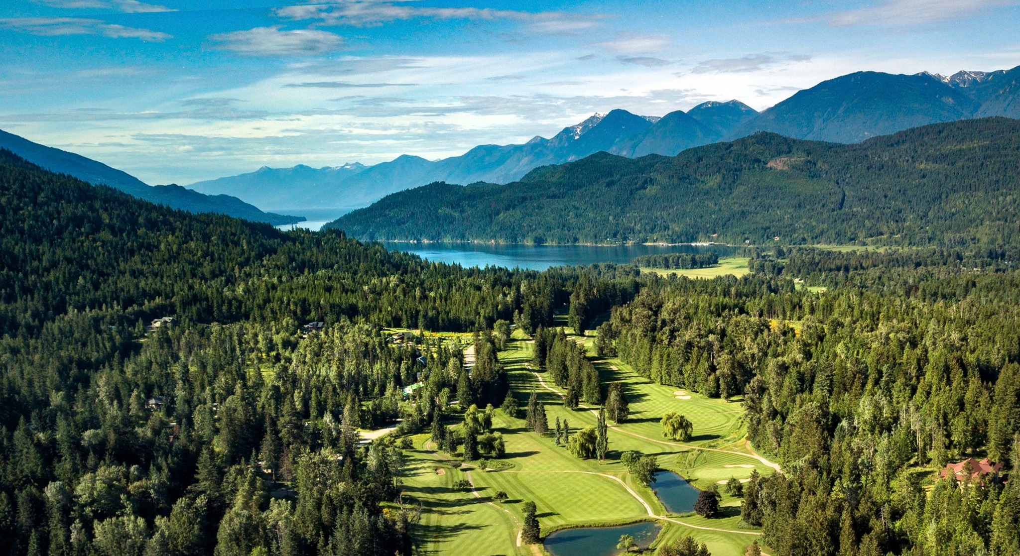 Aerial view of the Kokanee Springs Resort, showing fairway and greens set amongst trees, lake and mountains in far distance. 