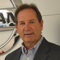 Jim Szakacs wears a suit jacket and stands in front of a large, silver Nissan symbol.