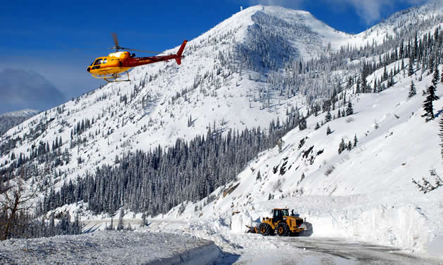 A helicopter hovers over an avalanche as a machine works to clear the road. A stunning mountain and blue sky are visible.
