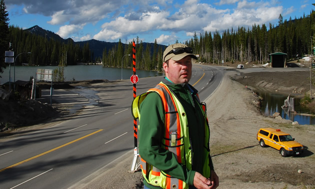 Cliff stands above a highway, lake, and green trees while wearing a safety vest.