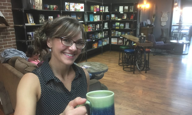 Candice Pickering, smiling into camera and holding up a cup of coffee. Bookstore in background. 