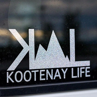 Close-up of white Kootenay Life sticker. Logo is stylized mountains sandwiched between backwards letter 'K' and the letter 'L', with the words 'Kootenay Life' underneath. 