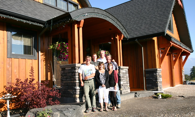 Group of seven people stand in bright sunlight in front of large timber house