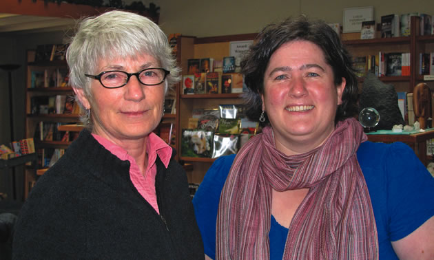 Silver-haired woman stands with younger, dark-haired woman before a wall of books on display. 