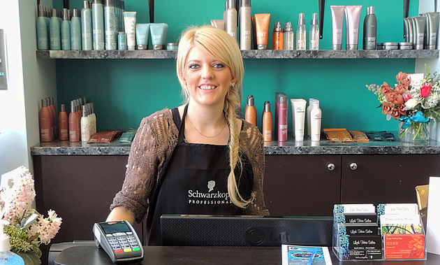 Sarah LeNeveu  stands behind the counter at Lysh Hair Salon in Fernie, B.C. Behind her is a turquoise wall and hair products. 