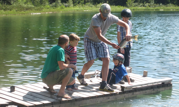 Two men—one holding a struggling fish—with three little boys on a dock on a sunny lake