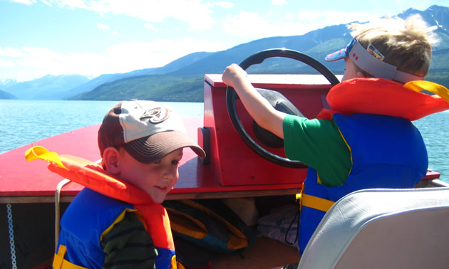 Close-up of two little boys wearing lifejackets, in a boat on a lake; one boy holds the steering wheel