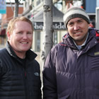 Two men wearing ski-jackets stand outdoors on a city street with mountains in the background