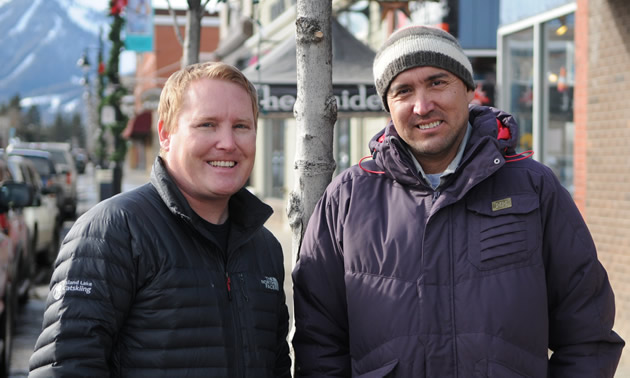 Two men wearing ski-jackets stand outdoors on a city street with mountains in the background