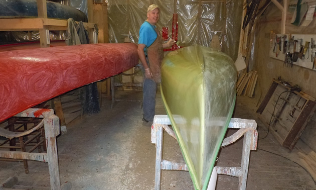 Photo of Mike Elliott working on a wooden canoe paddle with wood canoes behind