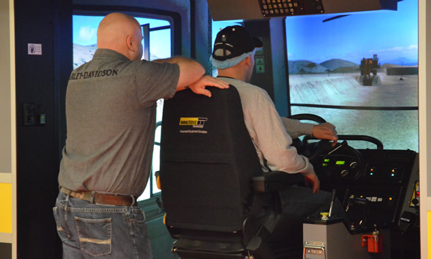 A man operates a haul truck simulator while another watches.