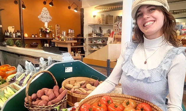 Smiling woman holding basket of tomatoes, other produce sitting on shelves in background, photo taken inside Morchella Market. 