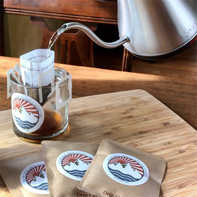 Hot water being poured over a single-serve coffee bag in a cup, with small packets of coffee bags in foreground of picture. 