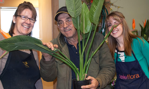 Lloyd holds and exotic flower and peeks out from between the leaves with Pat and employee Brittany on each side.
