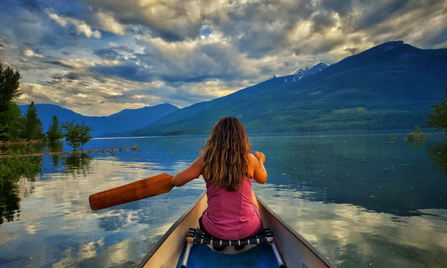 Young woman paddling a canoe, expansive lake and mountain views in background. 