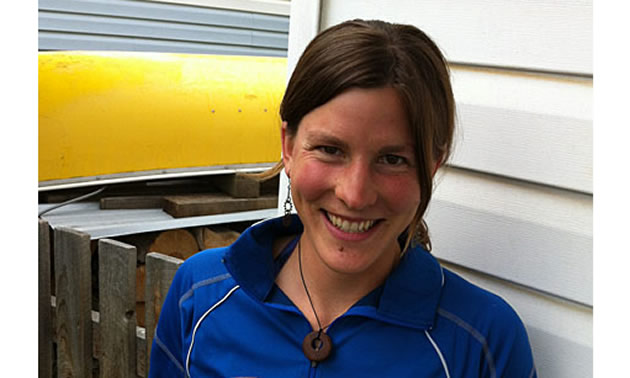 A brown haired young woman smiles at the camera. She wears a blue BC shirt and there is a yellow canoe in the background.