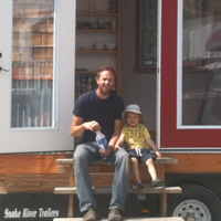 Man and young child sit on the  step in front of the open door of a tiny house