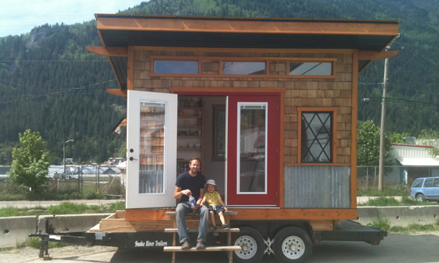 Man and young child sit on the  step in front of the open door of a tiny house