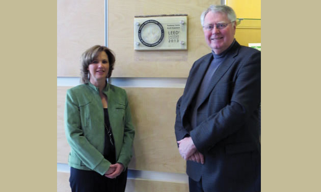 A woman in a green jacket and a tall man in a dark blue suit stand next to a LEED plaque. 