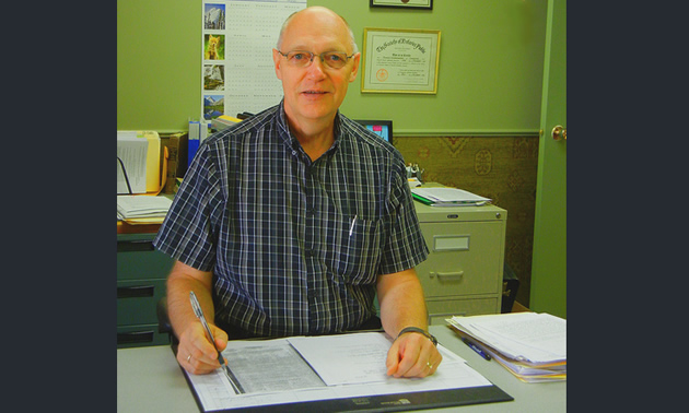 Ron Schatschneider sits behind a desk with a ledger and a pen.