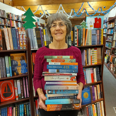 Letty Bartel, owner of Otter Books, stands in front of bookshelves holding a large stack of books. 