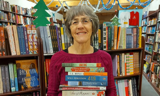 Letty Bartel, owner of Otter Books, stands in front of bookshelves holding a large stack of books. 