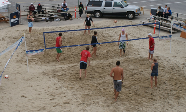A volleyball court is littered with players and is built of sand in a parking lot. 