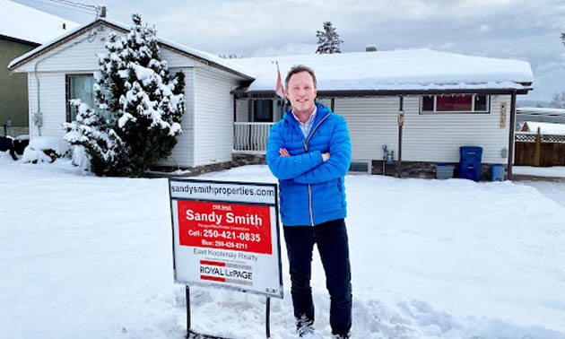 Phil Aston standing beside ‘For Sale’ sign in front of house on a snowy day. 