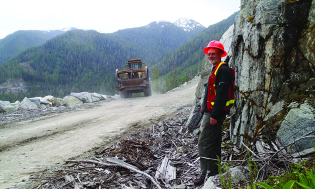 A worker stands next to a forestry road in the bush in British Columbia with heavy equipment in the background.