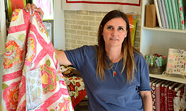Tammy Howard has long hair and a blue shirt. She is standing next to a pink and pastel flower quilt with bricks and shelving behind her.