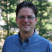 Dark-haired man wearing eyeglasses and a blue shirt; deciduous trees in the background