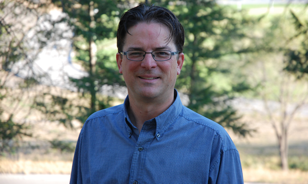 Dark-haired man wearing eyeglasses and a blue shirt; deciduous trees in the background