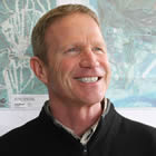 Rod Kessler, blonde and wearing a black jacket, smiles against a blue artwork background. 