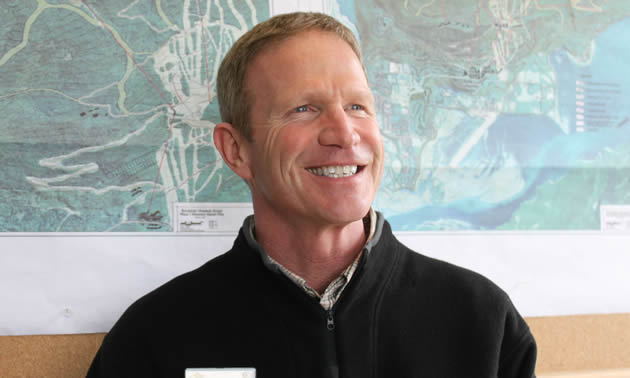 Rod Kessler, blonde and wearing a black jacket, smiles against a blue artwork background. 