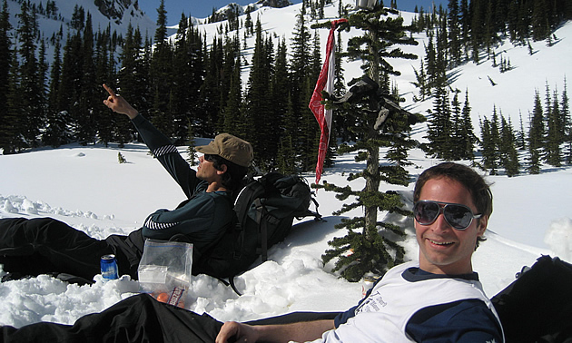 Two men in winter clothing are using backpacks as pillows while they take a break from a hike on the side of a snowy mountain slope.
