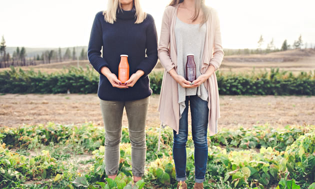 Natasha Benson (L) and Julie Taylor are holding two bottles of their cold-pressed juices while standing in a field.