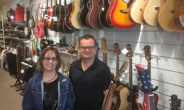 Jeremy and Felicity Youngward standing in their music store, with row of guitars behind them. 