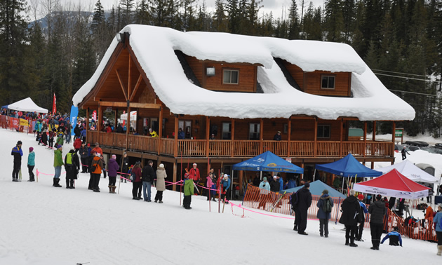 A snow covered and picturesque log cabin structure is surrounded by people and tents.