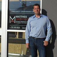 Robert wears a blue shirt and stands next to a logo on a window saying that the building was designed by Matthes Architecture.