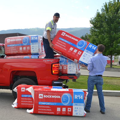 A Rockwool employee stands in the bed of a red Rockwool pickup truck, handing a bag of insulation to a Boundary region homeowner 