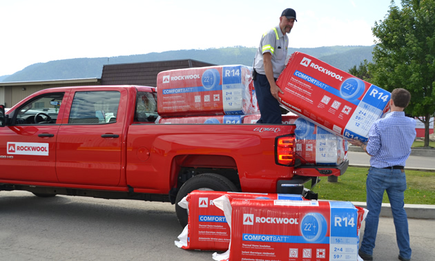 A Rockwool employee stands in the bed of a red Rockwool pickup truck, handing a bag of insulation to a Boundary region homeowner 