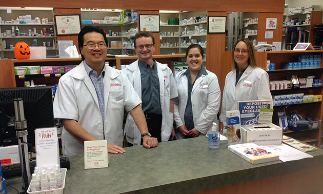 Four white-coated people, two men and two women, stand behind a counter in a pharmacy