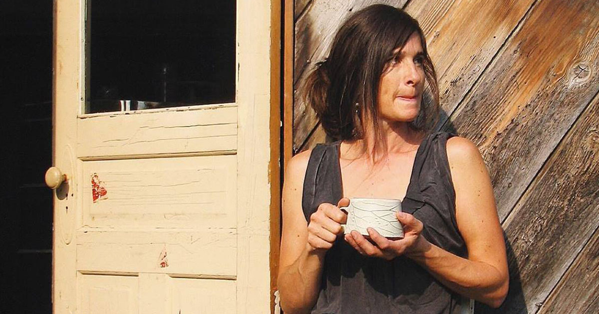 Sarah Pike sitting outside studio, holding mug and looking off into distance. 