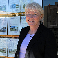 Shelley wears a black blazer and lilac shirt. She stands next to the door of her office with listings behind.