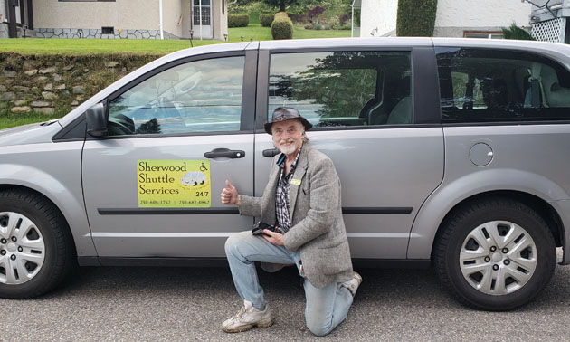 Mike Sherwood, owner of Sherwood Shuttle Services, kneeling beside his vehicle. 