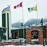 Three flags flying outside of a dark green and brick one-storey building, with a sign in the foreground saying District of Sparwood Municipal Office