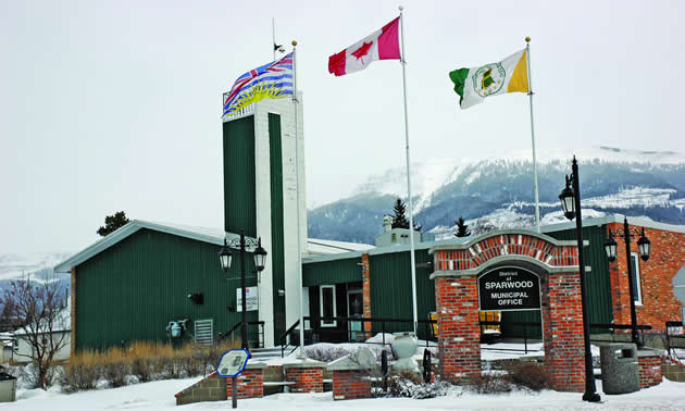 Three flags flying outside of a dark green and brick one-storey building, with a sign in the foreground saying District of Sparwood Municipal Office