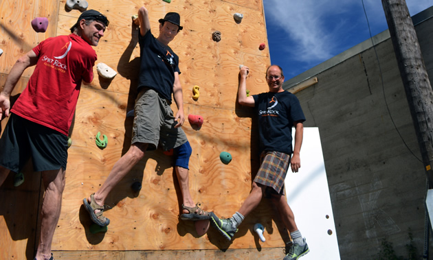 Three guys hold on to a rock-climbing wall with holds on plywood, and smile at the camera.