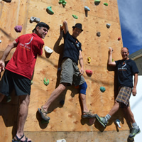 Three guys hold on to a rock-climbing wall with holds on plywood, and smile at the camera.