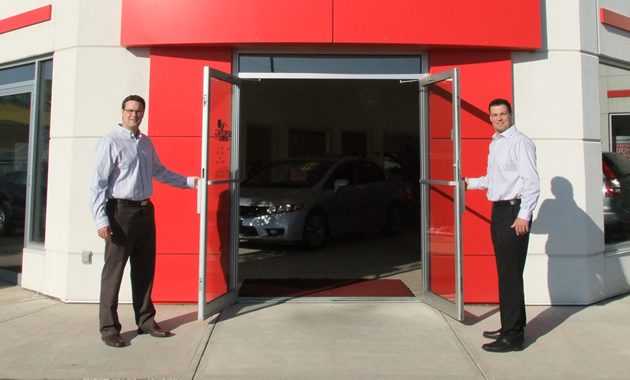 Two men stand at either side of a red entry emblazoned with the Honda symbol.